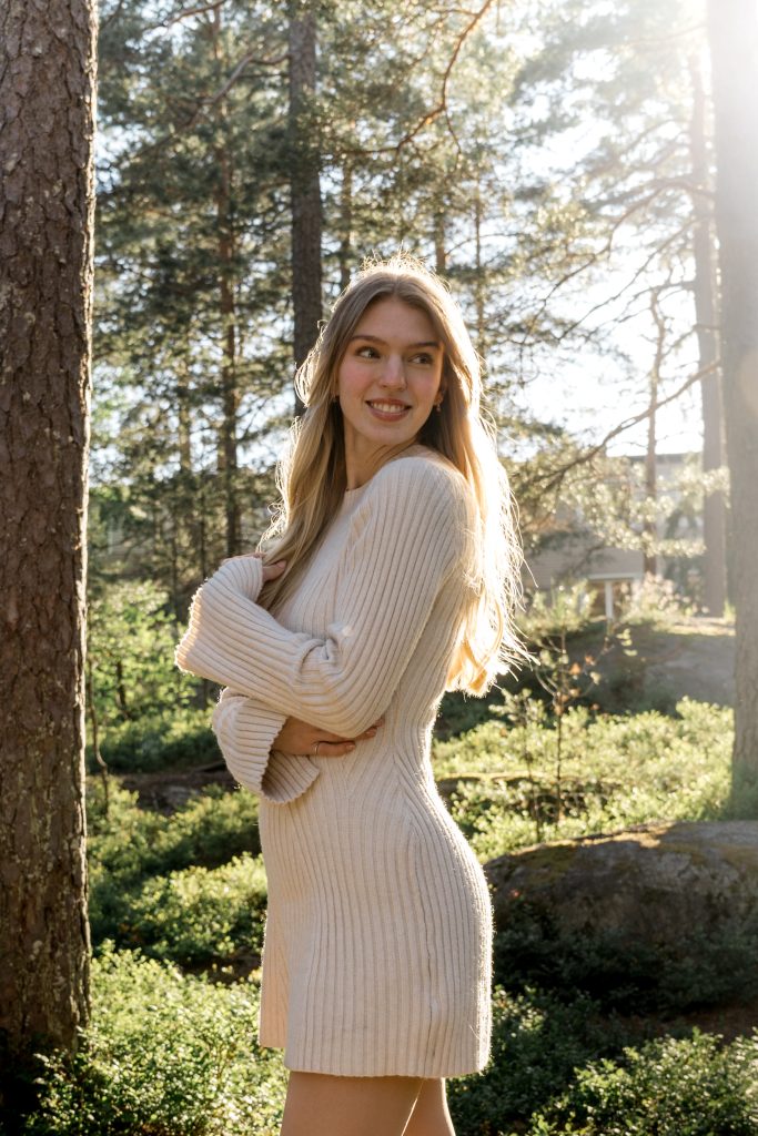 Portrett av en kvinne i en ribbet, kremfarget kjole som står i en solbelyst skog og smiler og ser til venstre for henne. Hun har langt blondt hår og er omgitt av trær og grøntområder, med sollys som filtrerer gjennom grenene i bakgrunnen.
