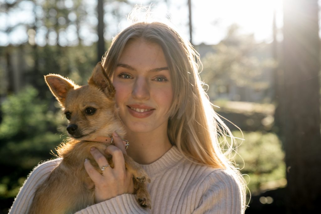 En kvinne med langt blondt hår smiler og holder en liten solbrun hund tett inntil ansiktet. De er utendørs i Oslo med sollys som filtrerer gjennom trær i bakgrunnen. Hun har på seg en lys kjole, og hunden har et rosa halsbånd, og lager et hjertevarmt portrett av hennes elskede kjæledyr.