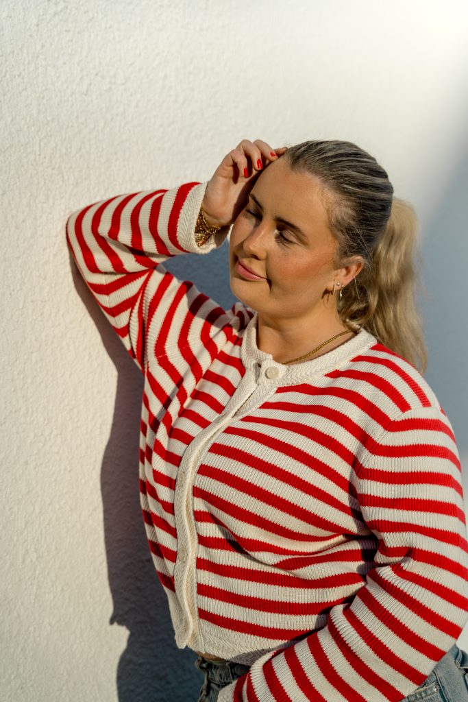 En kvinne står mot en hvit vegg, iført en rød og hvit stripet cardigan. Hun har øynene lukket, og høyre hånd berører pannen. Det blonde håret hennes er bundet tilbake i en hestehale, og hun smiler mykt. Sollys kaster en mild skygge.
