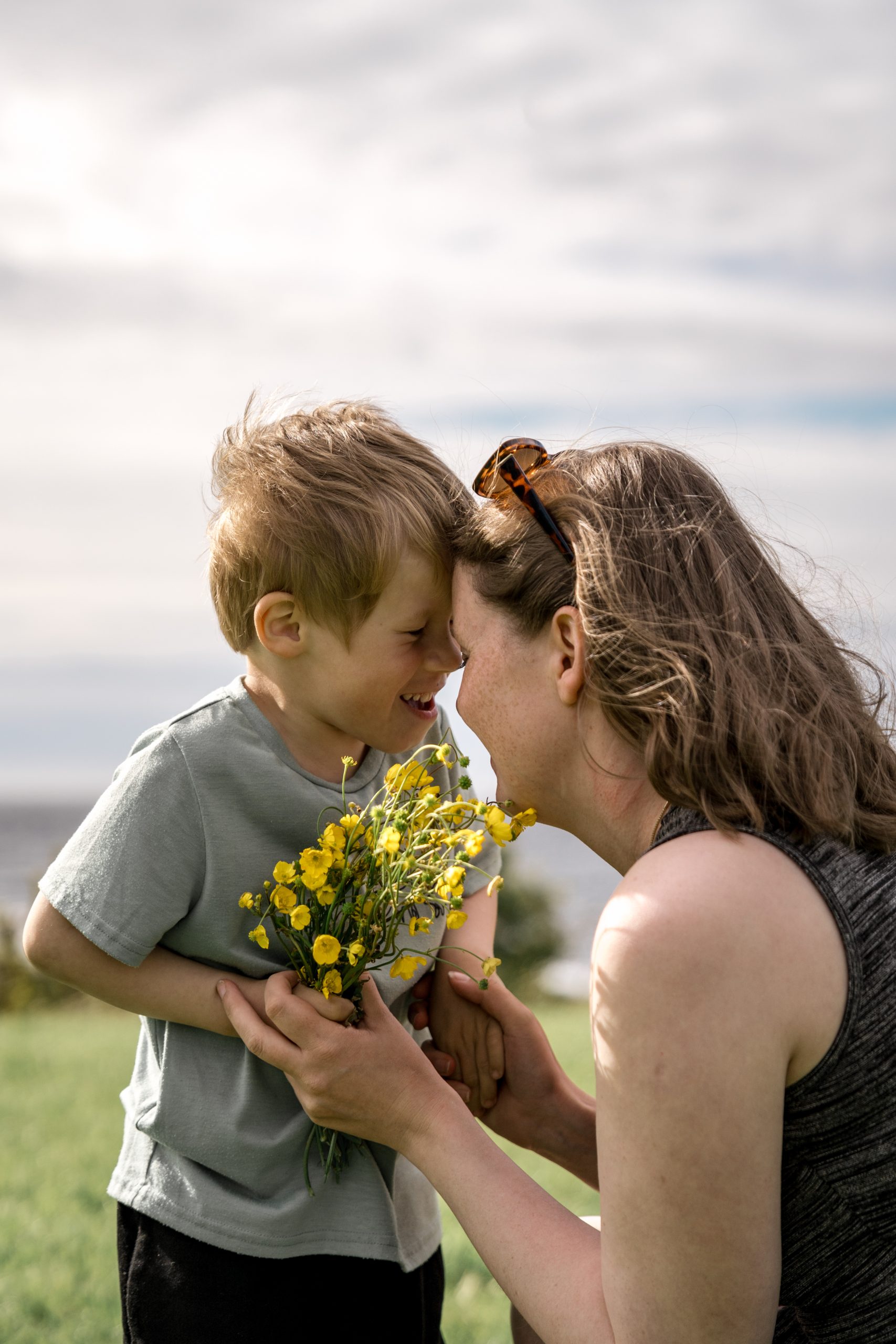 En mamma og en ung sønn deler et ømt øyeblikk utendørs. Gutten holder en bukett med gule blomster og lener pannen mot kvinnens panne. Kvinnen har solbriller på hodet. Bakgrunnen viser en gressmark under en delvis overskyet himmel.