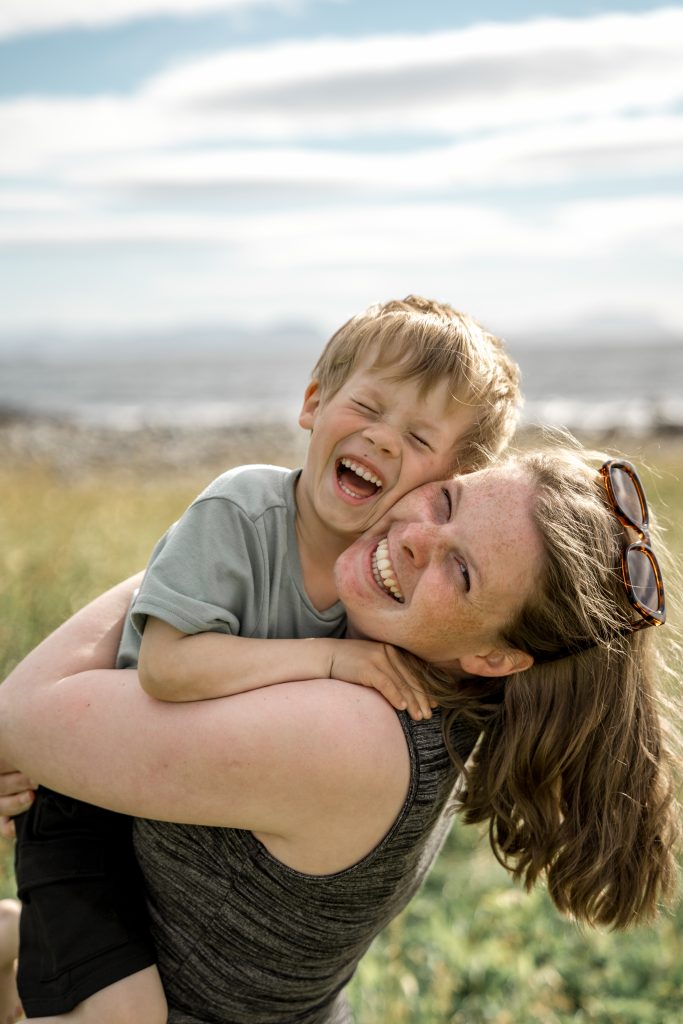En smilende kvinne med solbriller på hodet holder en lattermild ung gutt i armene mens hun står ute på en solrik dag. Bakgrunnen viser et uskarpt landskap med gress og en overskyet himmel, og fanger essensen av familiefotografering mens de nyter dette lekne, lykkelige øyeblikket sammen.