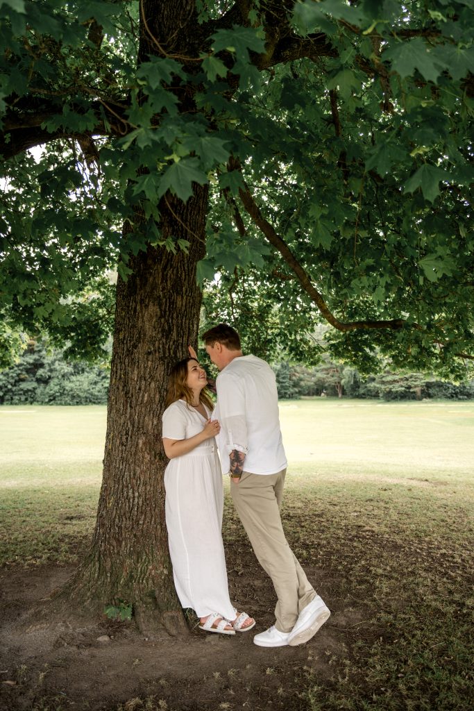 Et par står under et stort tre i en park. Kvinnen, iført en flytende hvit kjole og sandaler, stirrer opp på mannen. Mannen, kledd i hvit skjorte og beige bukser, lener seg mot treet og ser ned på henne. Det omkringliggende gresset er frodig og grønt.