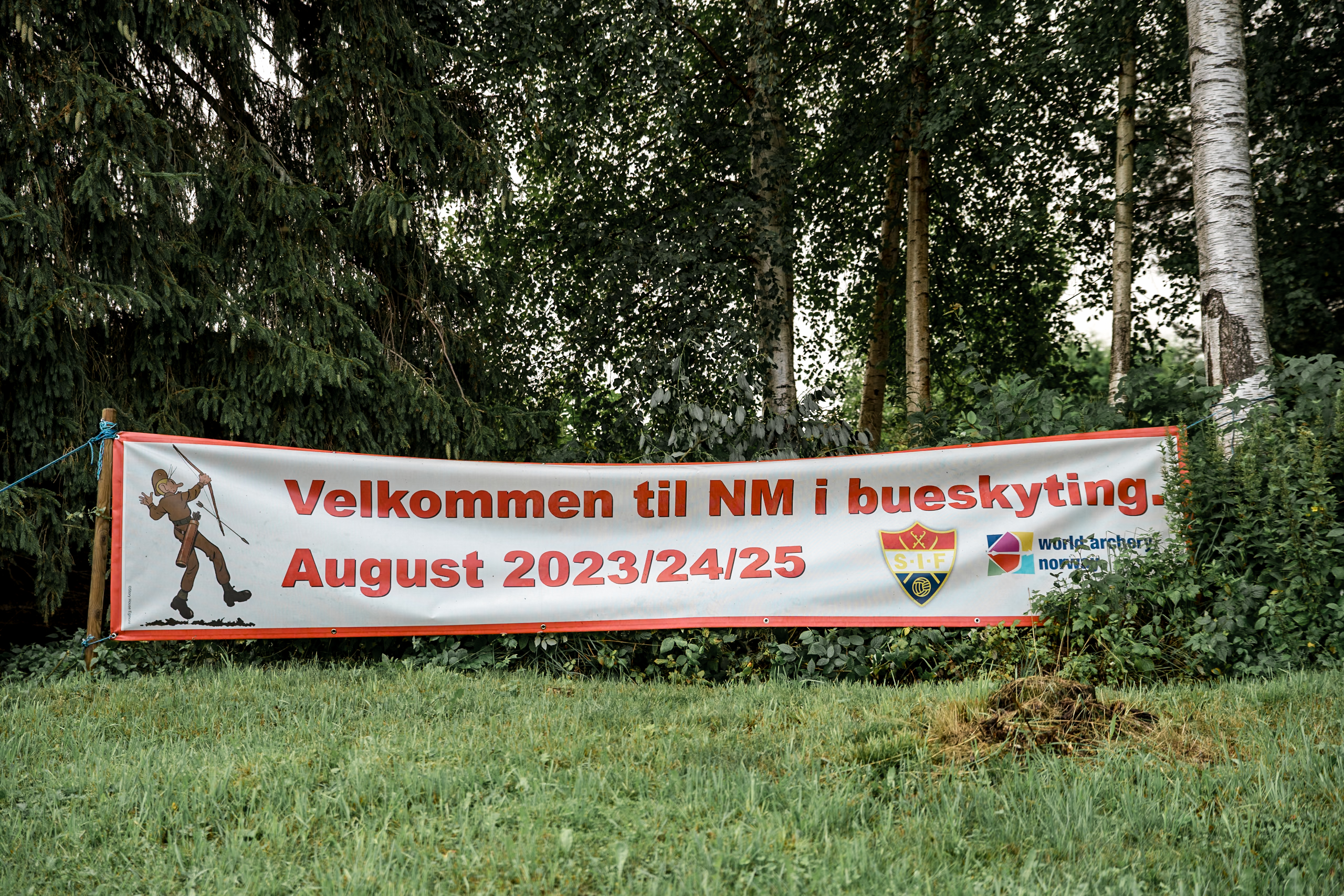 Et banner satt opp blant trær lyder «Velkommen til NM i bueskyting. August 2023/24/25». Banneret har logoer for Stomperudpila, Sørumsand Idrettsforening og Norges Burskytterforbund, og skaper et perfekt bakteppe for videre eventfoto.