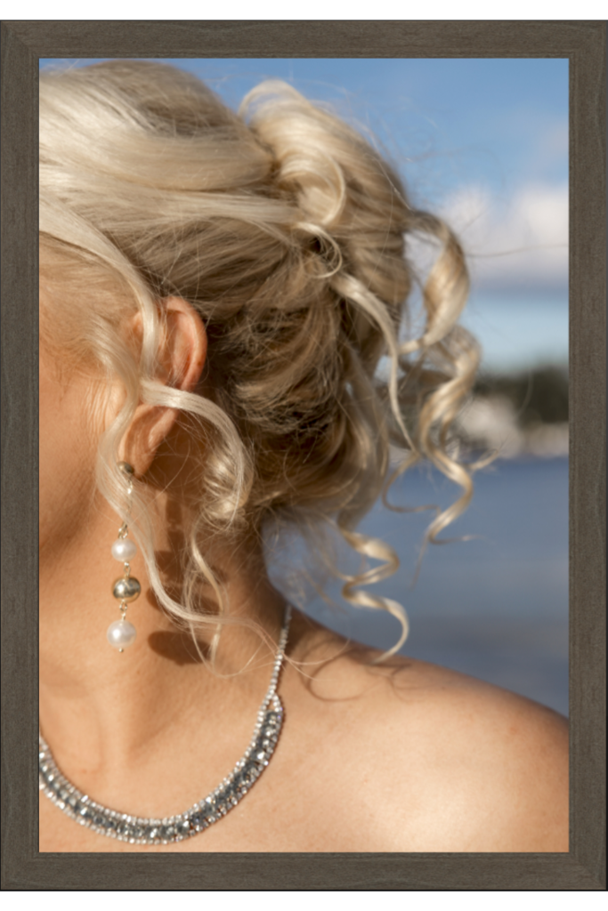 Et nærbilde av en kvinnes blonde hår stylet i en elegant updo med løse krøller, som minner om et trykkverdig mesterverk. Hun har på seg et glitrende kjede og en lang dinglende ørering, satt mot et uskarpt bakteppe av blå himmel og vann. Bildet er inni en treramme, noe som viser at det er et print.