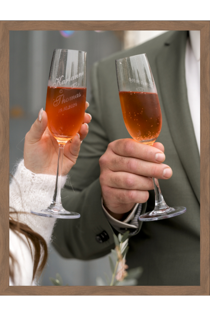 To personer skåler med personlige champagnefløyter fylt med en rosa drikke, og feirer et bryllup. Glassene, prydet med trykte navn og en dato, tilfører sjarm. Den ene har på seg en hvit genser og den andre en grønn dress, mens en treramme kunstferdig omgir scenen og viser at det er et print.