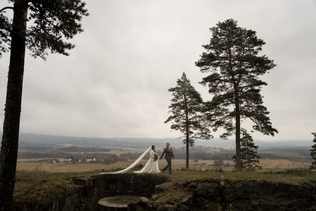 Et brudepar holder hender og går langs en gresskledd bakketopp, omfavnet av regnkysset høye furutrær. Bruden har på seg en lang hvit kjole med et flytende slør, som svaier forsiktig i vinden. Bakgrunnen har en overskyet himmel og vidstrakt utsikt over landskapet.