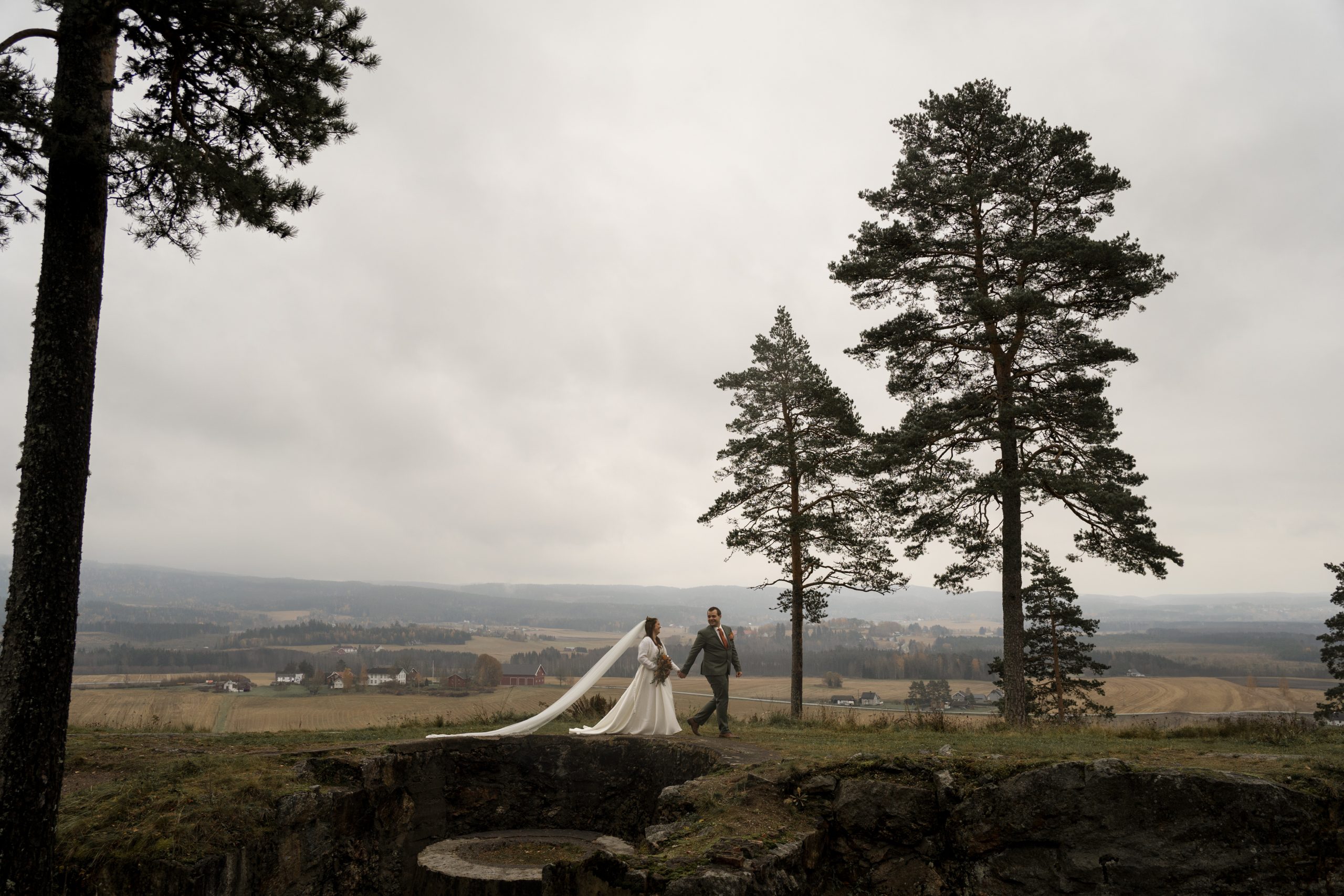 Et brudepar holder hender og går langs en gresskledd bakketopp, omfavnet av regnkysset høye furutrær. Bruden har på seg en lang hvit kjole med et flytende slør, som svaier forsiktig i vinden. Bakgrunnen har en overskyet himmel og vidstrakt utsikt over landskapet.