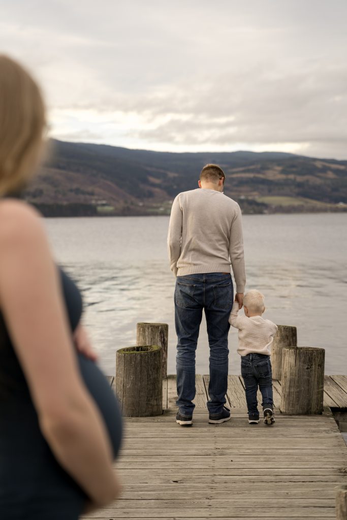 En gravid kvinne står i forgrunnen, litt ute av fokus, mens en mann og et lite barn holder hender og skuer over en innsjø fra en trebrygge, med fjell som strekker seg i bakgrunnen.