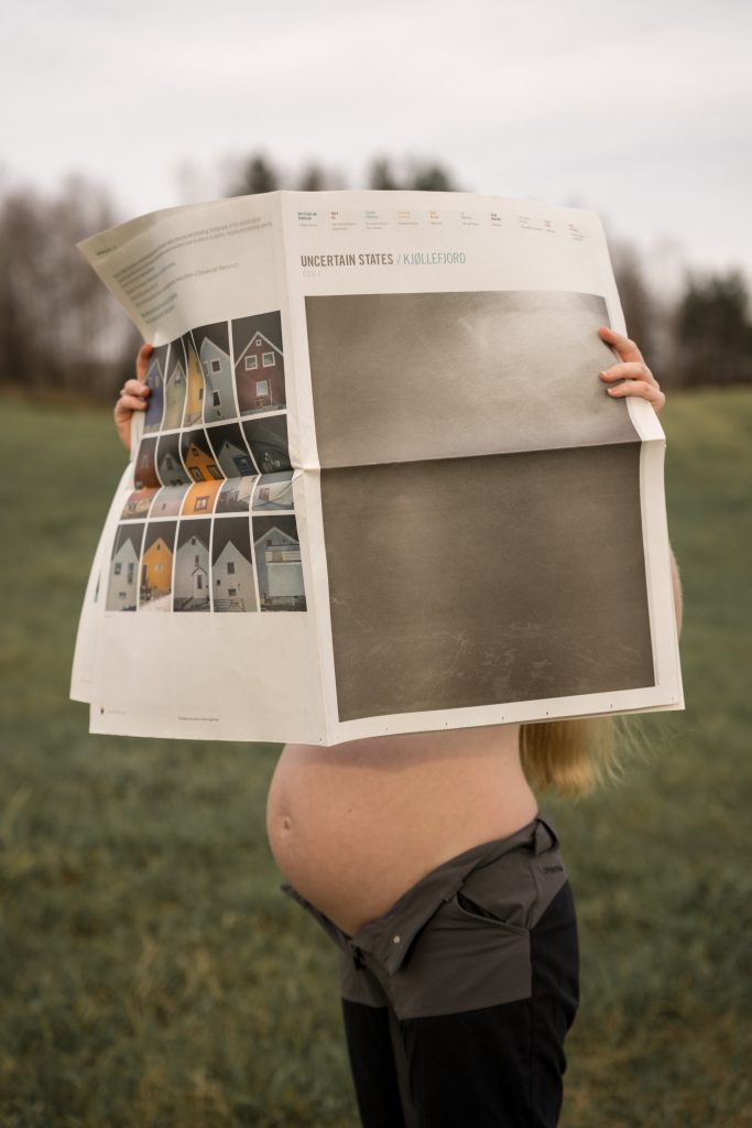 Et gravid individ står utendørs i en gressmark og holder en stor avis som skjuler ansiktet og overkroppen. Avisen viser bilder av hus på den ene siden og tekst på den andre. Himmelen er overskyet, med trær synlige i bakgrunnen, som antyder at regn som nærmer seg.