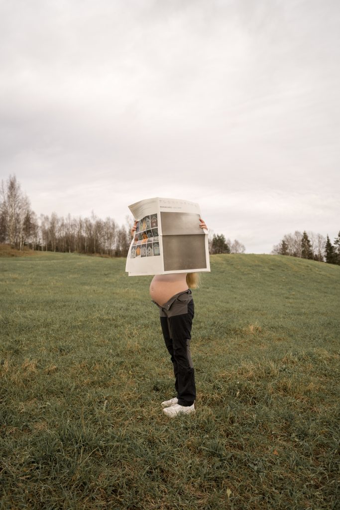 En gravid kvinne står i en gressmark med en stor avis som kun viser den gravide magen, midt i antydningen av forestående regn som er synlig på den overskyede himmelen, mens sparsomme trær rager stille i bakgrunnen.