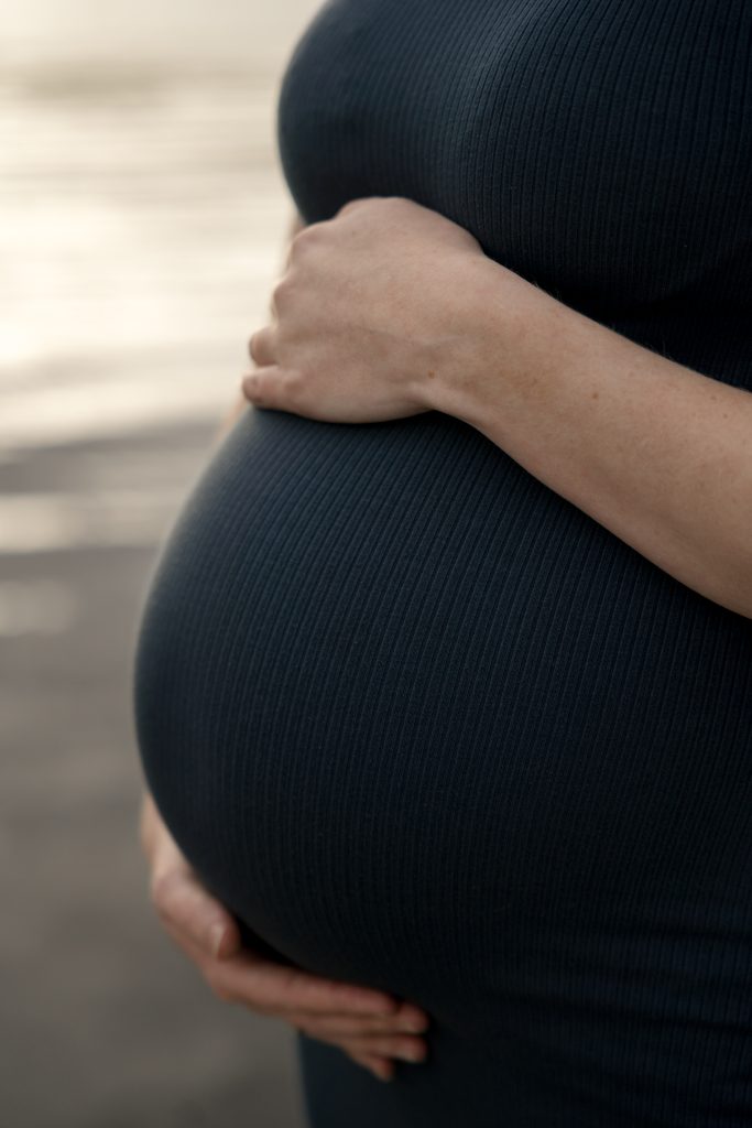 Et nærbilde av en gravid person i en ribbet, mørk kjole, som forsiktig vugger magen med én hånd over og én hånd under. Bakgrunnen er mykt uskarp.