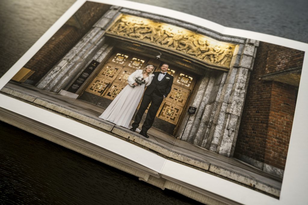 Et elegant bryllupsalbum ligger åpent på et bord, og viser et brudepar som smiler utenfor en utsmykket bygning med gyldne dører og intrikate utskjæringer over inngangen.