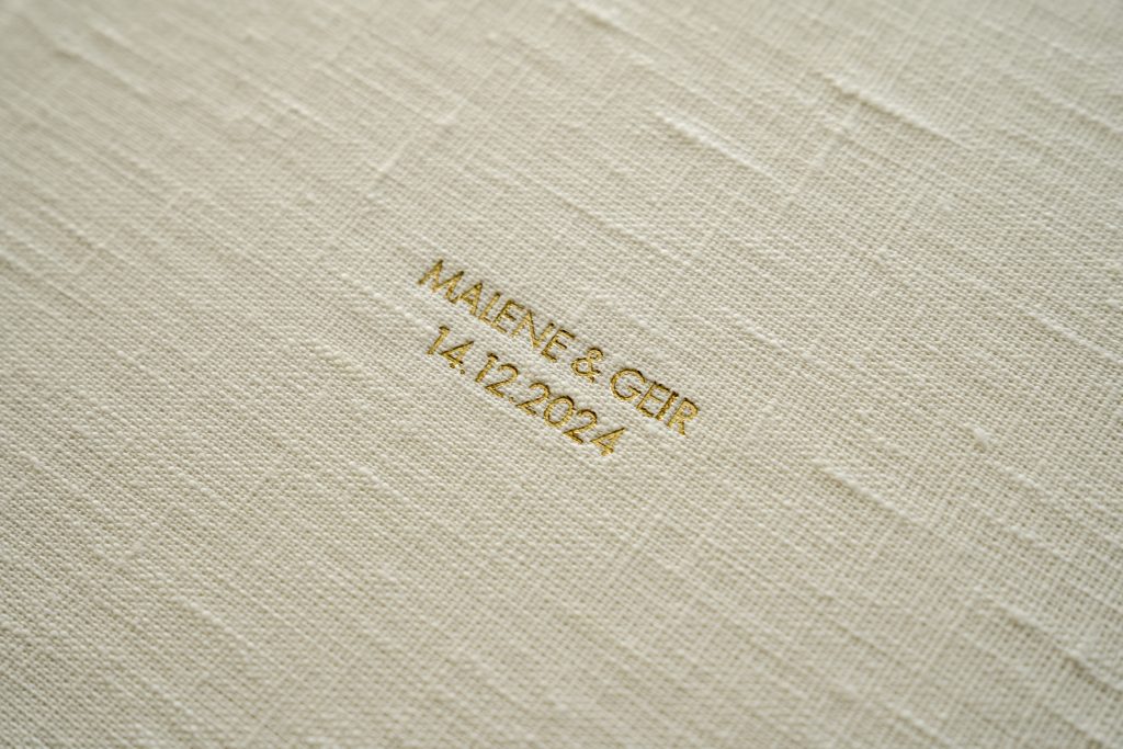 Nærbilde av albumomslag i teksturert kremstoff med gullpreget tekst med teksten "MALENE & GEIR 14.12.2024" i midten.