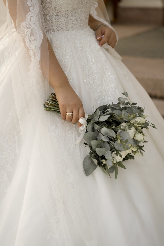 I et elegant bryllupsøyeblikk holder en brud i en hvit blondekjole en bukett med hvite blomster og grønt. Kjolen har intrikate blondedetaljer og slør med blonder. Hånden hennes, prydet med en ring, griper forsiktig buketten, som minner om ferdige bilder som fanger tidløs skjønnhet.