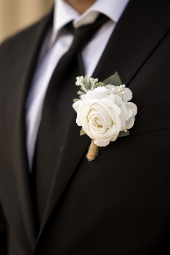 Et nærbilde fanger en person i en svart dress med en skarp hvit skjorte og svart slips, elegant utsmykket med en boutonniere av hvit rose og delikate blomster festet til jakkeslaget – perfekt egnet for ferdige bilder.