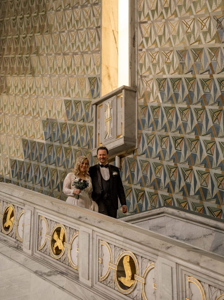 Et par i formelt antrekk går ned en marmortrapp ved Oslo Rådhus. Kvinnen holder en bukett, og begge smiler. Veggene har et geometrisk mønster med blå, gull og hvite farger. Bryllupsatmosfæren er elegant og sofistikert.
