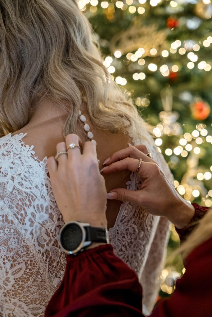 En hånd fester knapper på baksiden av en blondebrudekjole, satt i et koselig Lillestrøm-hjem med et pyntet juletre med lys og pynt i bakgrunnen. Bruden, som forbereder seg til bryllupsdagen, har langt, bølget blondt hår som faller nedover skuldrene.