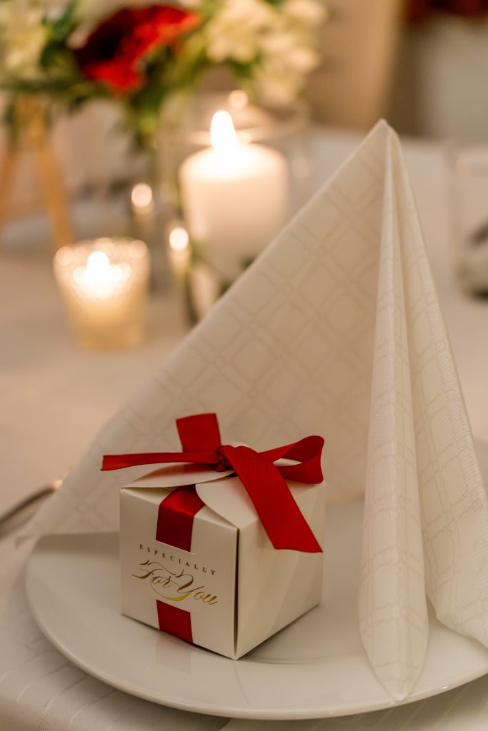 En hvit gaveeske med rødt bånd hviler på en hvit tallerken ved siden av en elegant brettet serviett. I bakgrunnen bidrar tente stearinlys og uklare blomster til den varme, festlige atmosfæren til et Lillestrøm-bryllup.