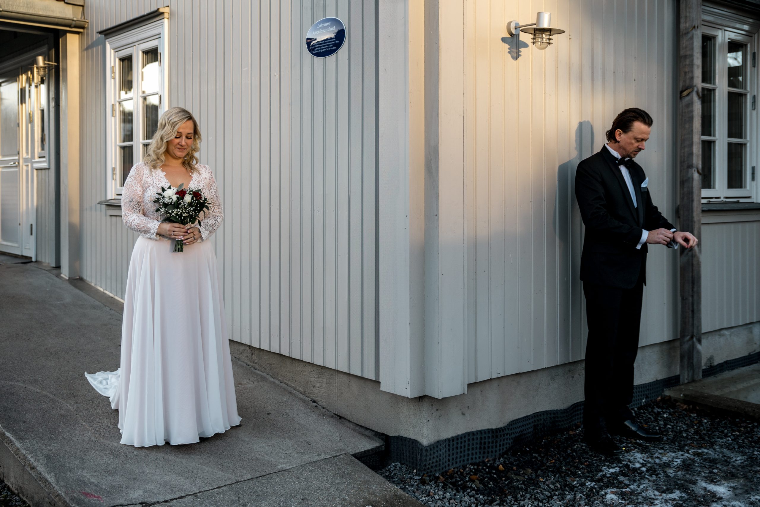 I Lillestrøm holder en brud i hvit kjole en bukett på verandaen, klar til bryllupet. Rundt hjørnet ser brudgommen i den svarte smokingen ivrig på klokken for deres first look. Verandaen er en del av en sjarmerende lys trebygning.