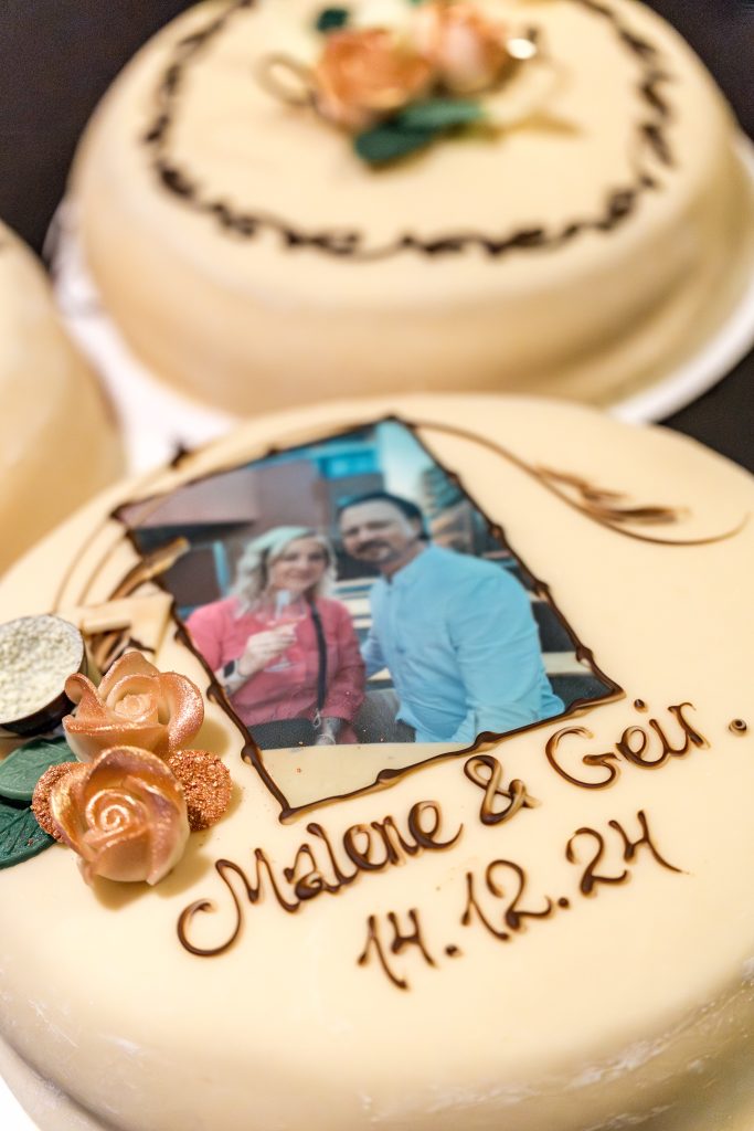 Nærbilde av en fantastisk bryllupskake med et bilde av paret. Gylne roser pryder designet, med "Malene & Geir 14.12.24" elegant skrevet i sjokolade. I bakteppet bidrar Lillestrøm-inspirerte kaker til den fortryllende fremvisningen.