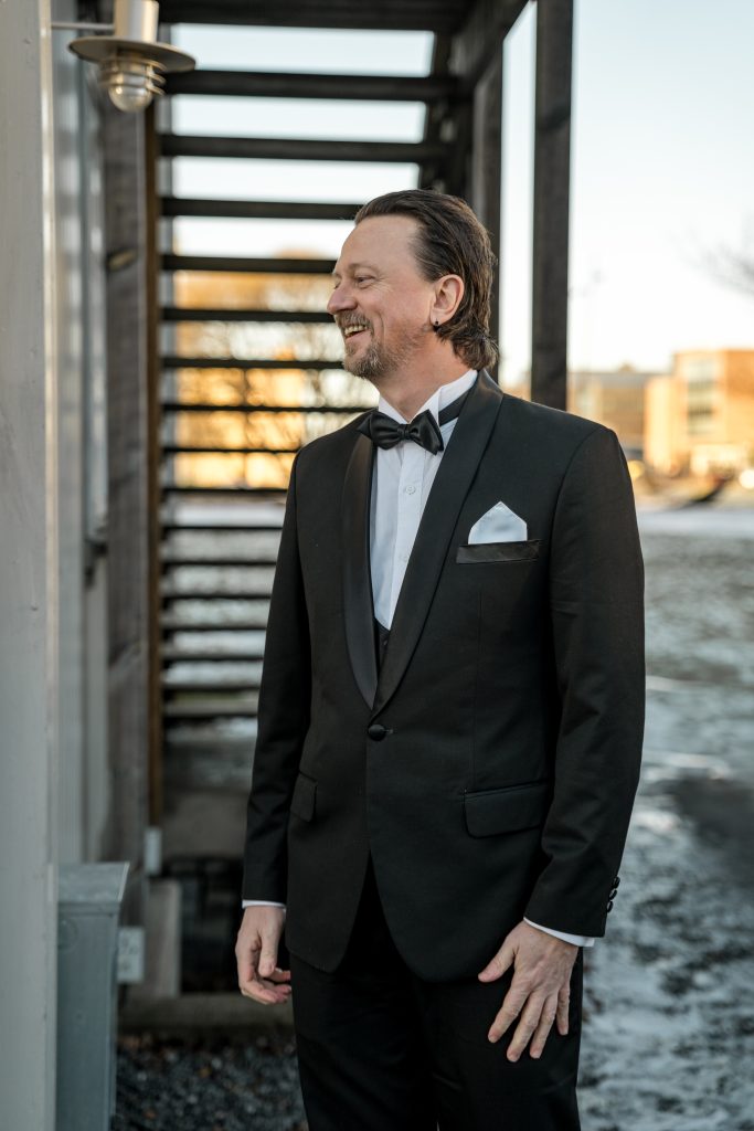 En mann i svart smoking med hvit skjorte og sløyfe står utenfor og smiler på bryllupsdagen. Han befinner seg i nærheten av et bygg med metalltrapp i Lillestrøm. Bakken er snørik, og bakgrunnen er uskarp. Han er i ferd med å se bruden i en first look.