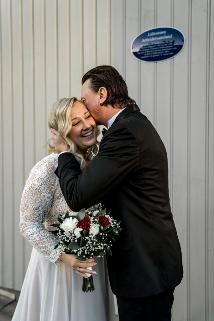 En smilende brud i en hvit blondekjole holder en bukett med røde og hvite blomster. Brudgommen i mørk dress kysser pannen hennes og fanger et perfekt bryllupsøyeblikk. De står foran en lysegrå vegg med blå plakett, som minner om sjarmerende Lillestrøm-scener.