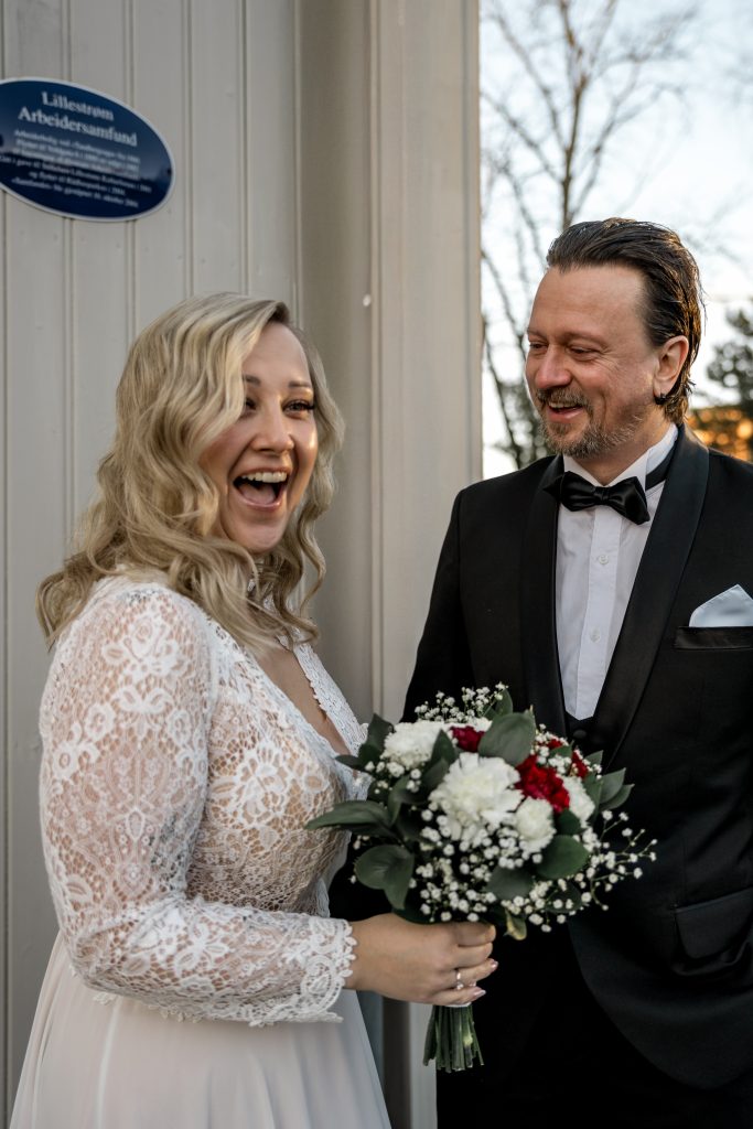 En brud i blondekjole stråler mens hun holder en bukett med hvite og røde blomster. Ved siden av henne smiler en mann i svart smoking. De står i nærheten av en bygning med blå plakett, som antyder Lillestrøms sjarmerende gater.