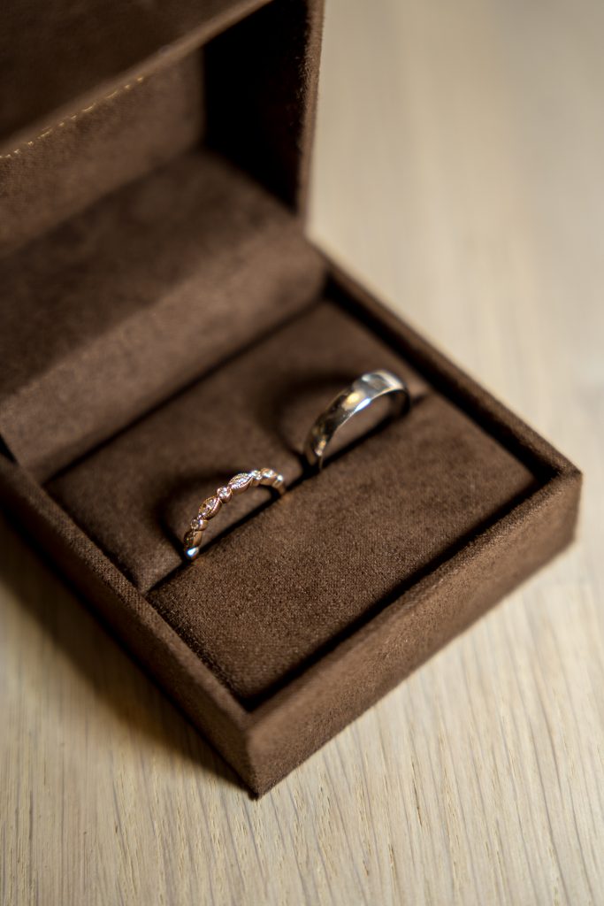 Et par ringer inne i en åpen brun fløyelsringboks, perfekt for et bryllup. Den ene ringen er belagt med små diamanter, mens den andre er et enkelt, polert bånd. Boksen hviler på en lys treflate i koselige Lillestrøm-omgivelser.