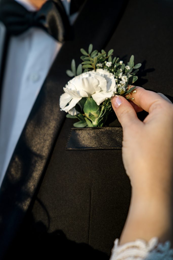 Ved et bryllup fester brudens hånd forsiktig en boutonniere med hvit blomst og grønt på en elegant svart dressjakke. Individet er kledd i en hvit skjorte og supplert med en klassisk svart sløyfe.