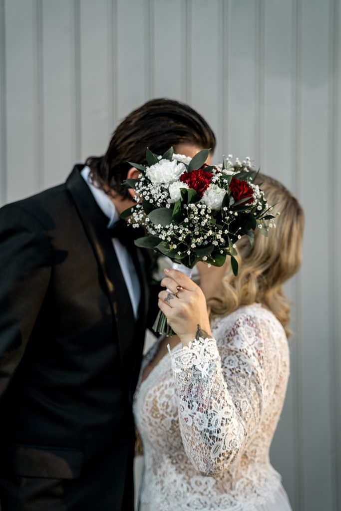 Et par i bryllupsantrekk, som feirer bryllupet sitt, holder en bukett foran ansiktene sine. Buketten viser røde og hvite blomster med grønt. Bruden har på seg en blondekjole, og brudgommen har på seg en svart dress mot en lysegrå panelbakgrunn som minner om Lillestrøms sjarm.