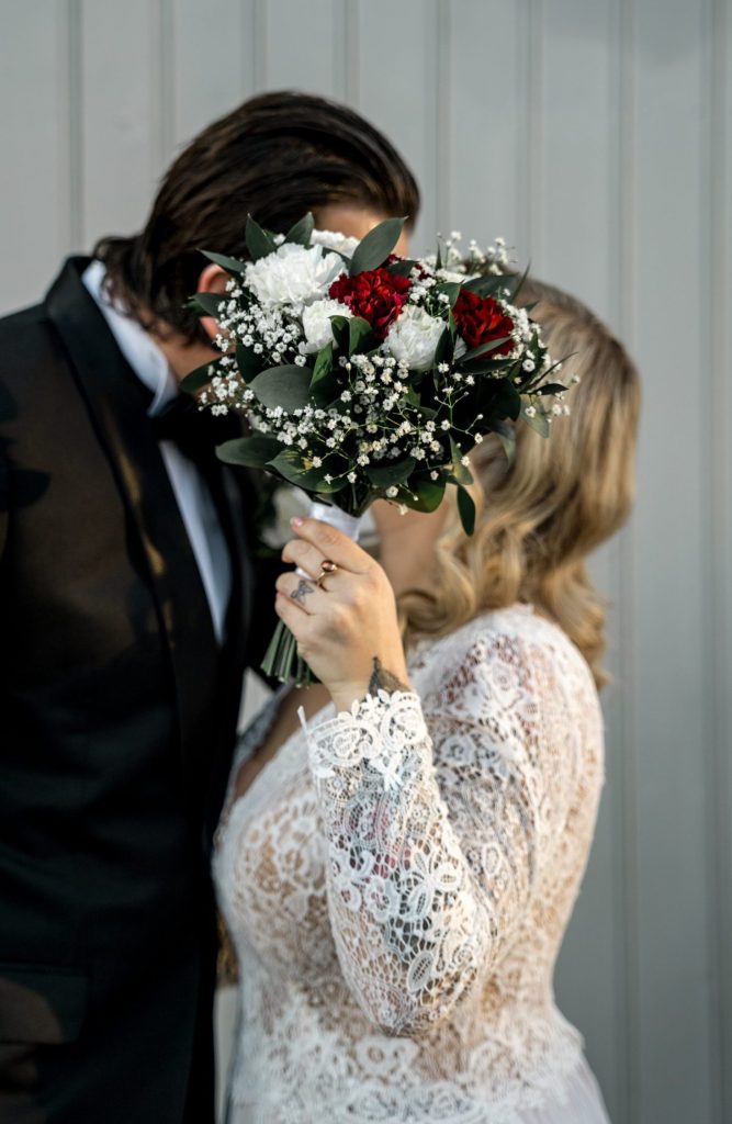 Et par i bryllupsantrekk, som feirer bryllupet sitt, holder en bukett foran ansiktene sine. Buketten viser røde og hvite blomster med grønt. Bruden har på seg en blondekjole, og brudgommen har på seg en svart dress mot en lysegrå panelbakgrunn som minner om Lillestrøms sjarm.