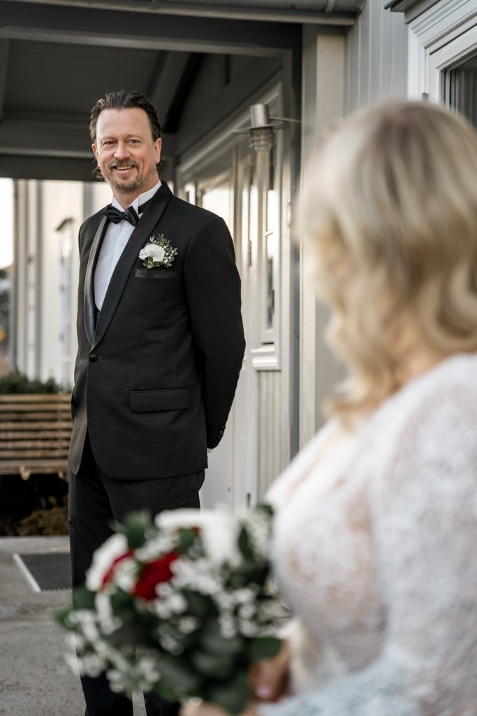 En mann i smoking med boutonniere står utenfor en bygning i Lillestrøm og smiler til bruden i forgrunnen som holder en bukett med røde og hvite blomster. Scenen antyder en bryllupsfest.