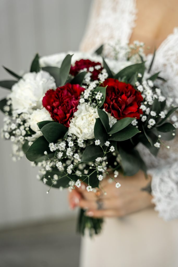 I en blondekjole holder en brud en bukett med røde og hvite blomster, omgitt av frodige grønne blader, perfekt for et vinterbryllup i Lillestrøm.