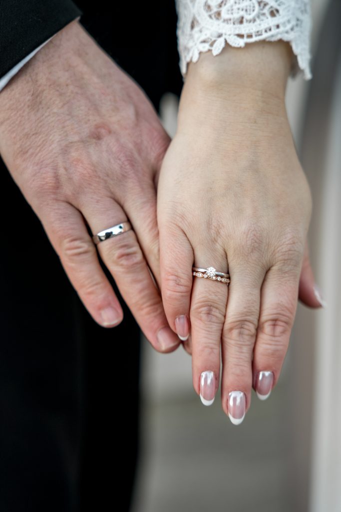I et ømt øyeblikk under et bryllup i Lillestrøm berører to hender forsiktig: hans utsmykket med et sølvbryllupsring og hennes med en diamantforlovelsesring og bryllupsring, det blondekantede ermet tilfører eleganse mot det myke, uskarpe bakteppet.
