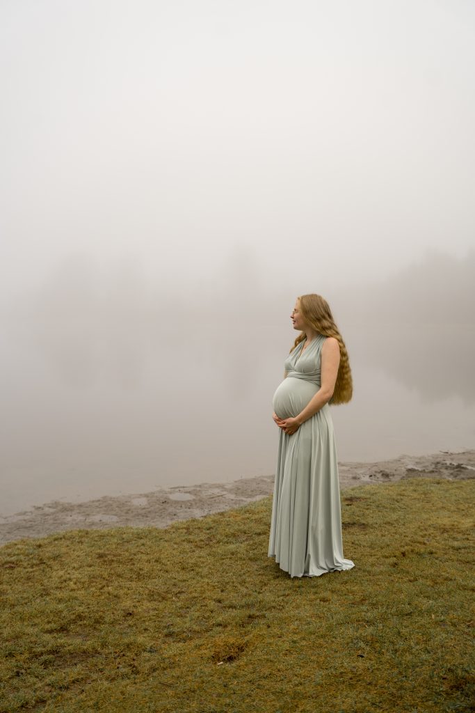 En gravid kvinne i en lang, flytende lysegrønn kjole står på gressmark ved en tåkete innsjø, og legemliggjør skjønnheten til gravidbilder ute. Den tåkete bakgrunnen skjuler trær og skaper en rolig og rolig atmosfære.