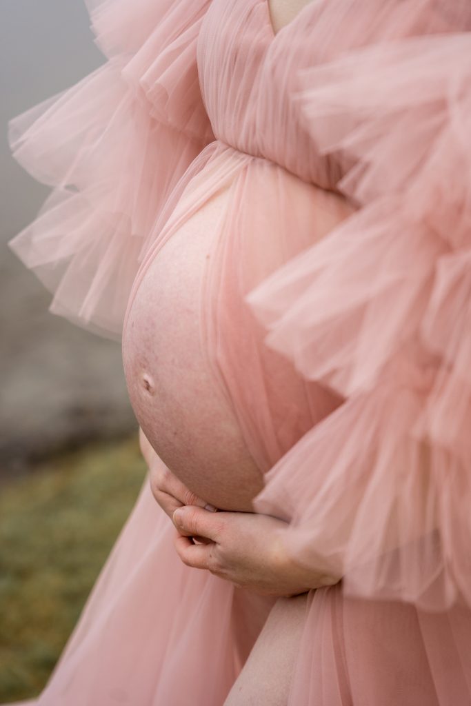 Et nærbilde av en gravid kvinne i en rosa, rynket kjole som forsiktig vugger den nakne magen med begge hender. Det myke stoffet og utendørsmiljøet skaper en delikat, rolig atmosfære.