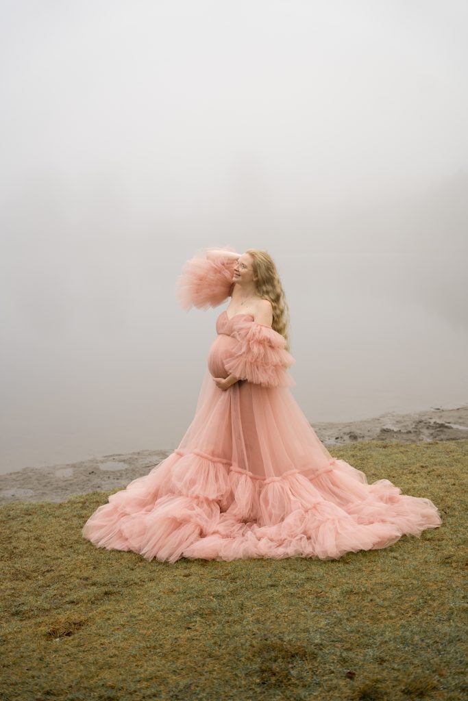 En kvinne i en flytende, krøllet rosa kjole står på gresset nær en tåkete innsjø. Den tåkete bakgrunnen skjuler trærne og vannet, og skaper en drømmende, eterisk atmosfære på dette gravidbildet.