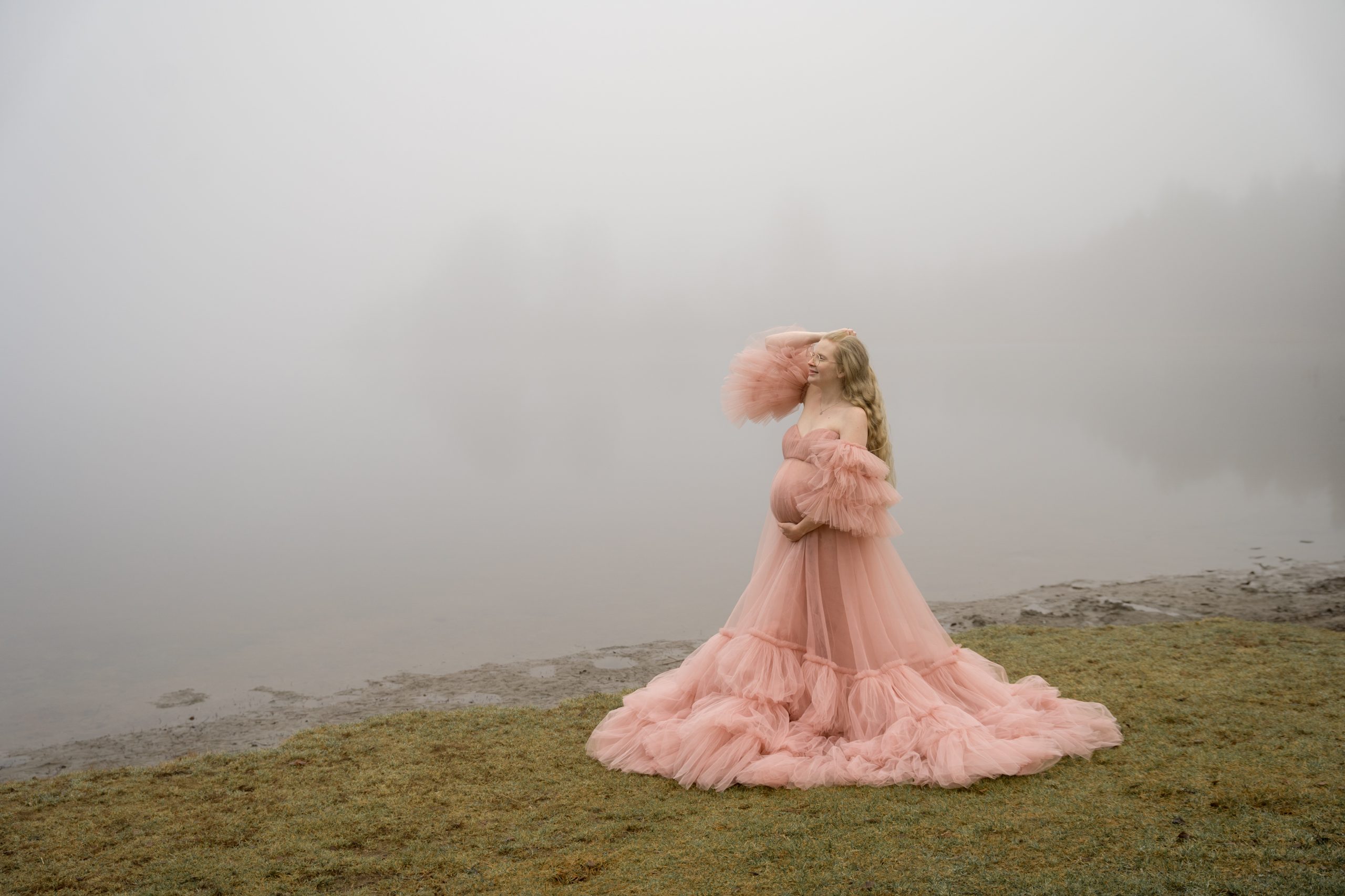 En kvinne i en flytende, krøllet rosa kjole står nær en tåkete innsjø, og drar hånden gjennom håret. Denne gravidbilder-scenen har tåkete trær og vann, og skaper en drømmende, eterisk atmosfære.