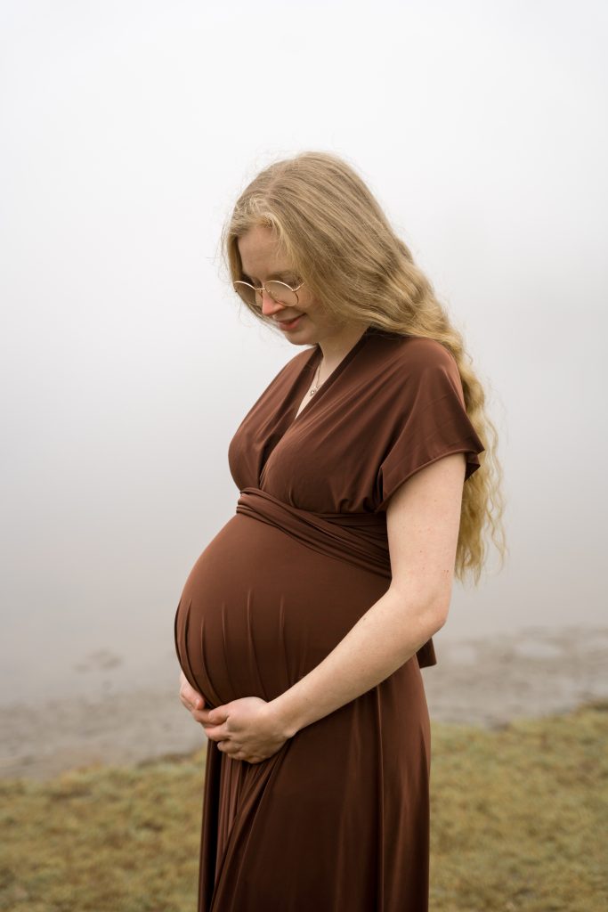 En gravid kvinne med langt blondt hår, iført briller og en brun kjole, står på gresset i en tåkete, uskarp bakgrunn — gravidbilder ute stil — og vugger forsiktig med magen og smiler mens hun ser ned.
