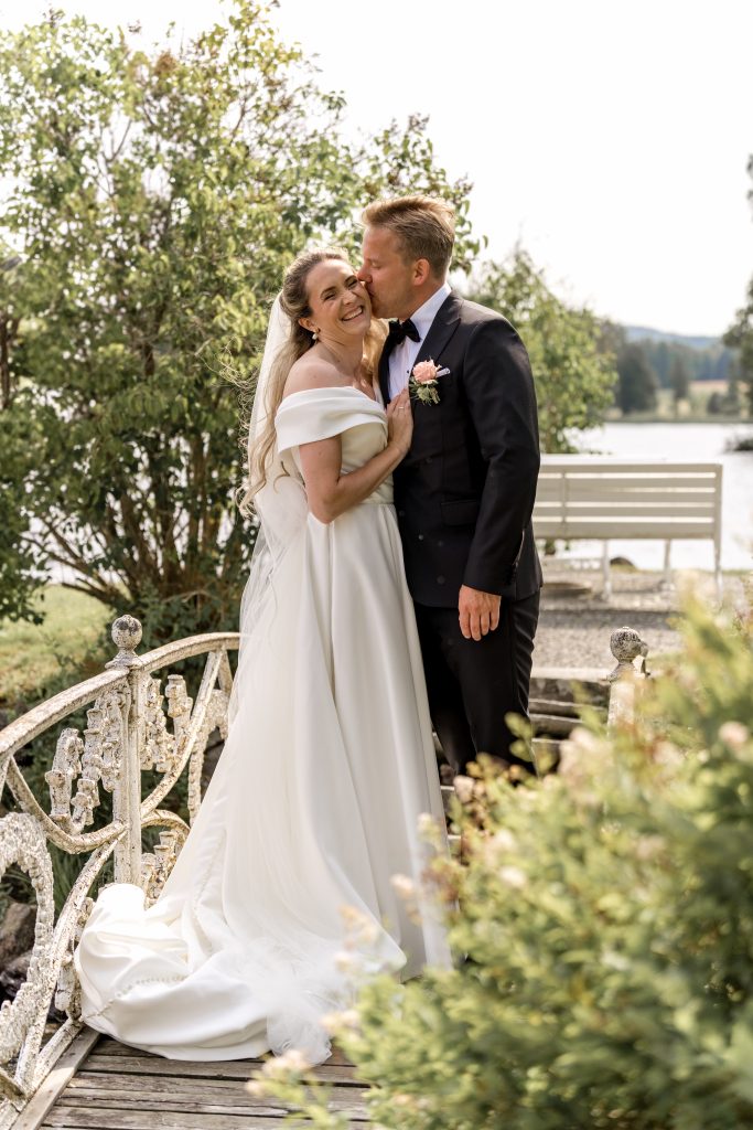 Brud smiler mens brudgom kysser henne på kinnet i hagen
