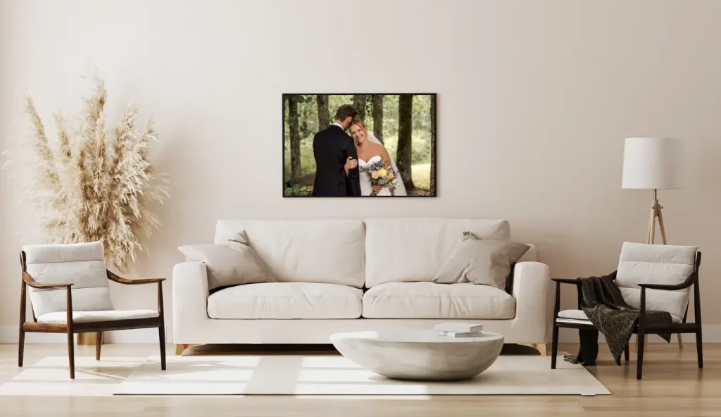 En nøytral stue med en hvit sofa, to lenestoler, et rundt salongbord, gulvlampe og dekorativt pampasgress. På veggen over sofaen henger et innrammet bryllupsfoto av et par.